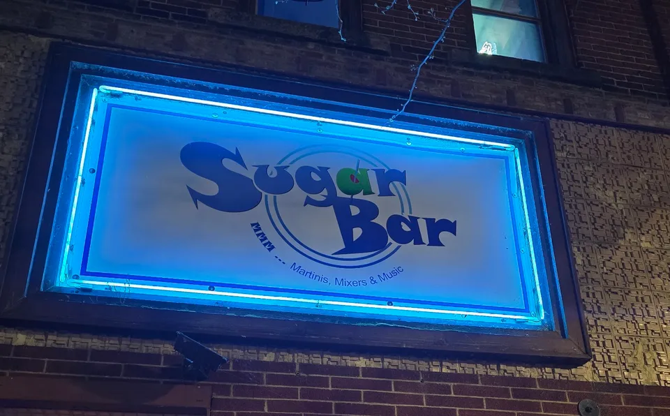 The illuminated Sugar Bar sign at night above the brick storefront at 835 Main St, reading MMM ... Martinis, Mixers & Music